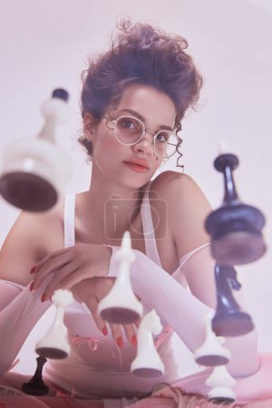 Photo for Beautiful young woman with chess pieces around, retro glasses and thoughtful expression. Promotion of a womens chess tournament. Concept of beauty and fashion, vintage, boudoir style - Royalty Free Image
