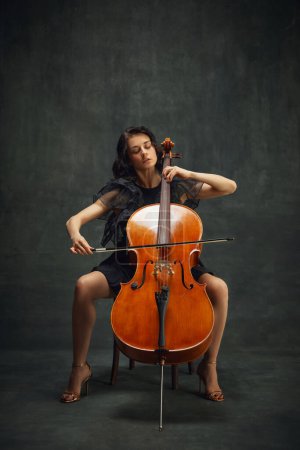 Elegant, passionate musician, beautiful woman in black dress sitting and playing cello against dark green vintage background. Concept of classical art, retro style, music, inspiration, orchestra event