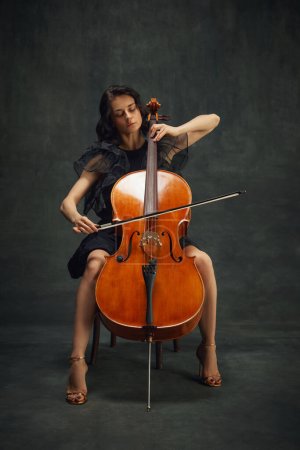 Mujer joven tierna, violonchelista tocando el violonchelo, silla sentada sobre fondo vintage verde oscuro. Cartel del concierto de música clásica. Concepto de arte clásico, estilo retro, música, inspiración