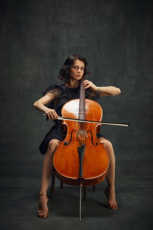 Beautiful young woman, cellist sitting on chair with cello, looking at camera on vintage green background. Cultural events with classic music performances. Concept of classical art, retro style, music