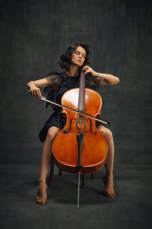 Passionate young woman, musician sitting with eyes closed and playing cello over vintage green background. Being fully into melody. Concept of classical art, retro style, music, inspiration