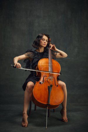 Cellists debut, classical album. Elegant young woman making solo performance, playing cello against dark vintage green background. Concept of classical art, retro style, music, inspiration