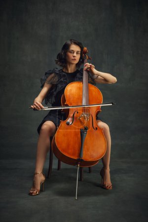 Beautiful young woman, cellist sitting with cello on vintage green background. Magazine cover about talented musicians in the classical music scene. Concept of classical art, retro style, music