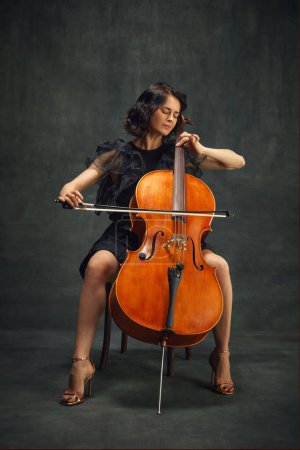 Cellist, elegant woman in deep concentration, playing cello passionately with eyes closed against vintage green background. Concept of classical art, retro style, music, inspiration
