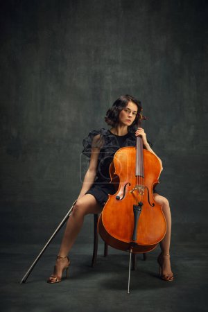 Elégant violoncelliste féminin assis sur une chaise avec violoncelle, regardant la caméra sur fond vert vintage. Programme de musicothérapie classique. Concept d'art classique, style rétro, musique, inspiration