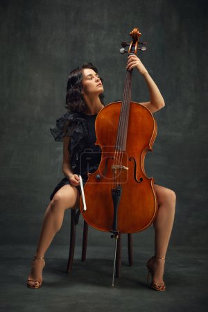 Elegant, beautiful young woman, cellist sitting with wooden cello against dark green vintage background. Acoustic concert. Concept of classical art, retro style, music, inspiration