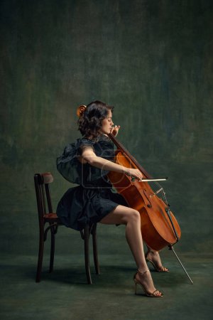 Attractive, elegant young woman, cellist, musician in black dress sitting on chair and playing cello against dark vintage background. Concept of classical art, retro style, music, inspiration