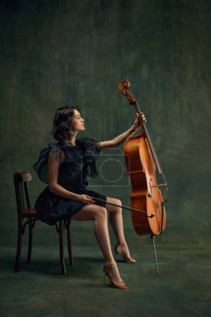 Classical music live performance. Elegant young woman, cellist, passionate musician sitting with cello against dark vintage green background. Concept of classical art, retro style, music, inspiration