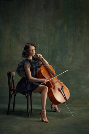 Pensive female musician, cellist sitting on chair with cello, in thoughtful pose on vintage green background. Poster for orchestras seasonal performances. Concept of classical art, retro style, music