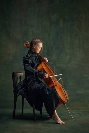 Tender young woman in black dress, cellist sitting on chair and playing cello over dark green background. Combination of elegance and music. Concept of classical art, retro style, music, inspiration