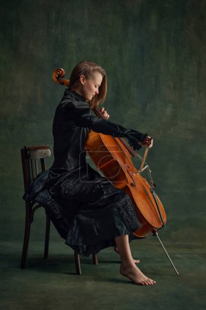 Passionate musician, elegant young woman, cellist in black dress sitting on chair and playing cello against dark vintage background. Concept of classical art, retro style, music, inspiration