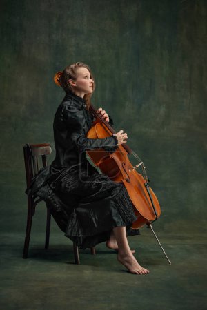 Elegante mujer joven, talentosa, violonchelista apasionada mirando hacia arriba, reflejando creatividad e inspiración mientras toca. Tomando inspiración. Concepto de arte clásico, estilo retro, música, inspiración