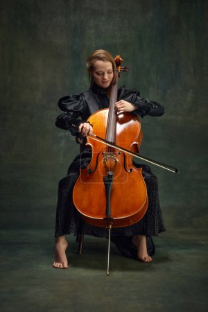 Beautiful young woman, cellist sitting on chair and playing cello against vintage green background. Classic music performances. Concept of classical art, retro style, music, inspiration