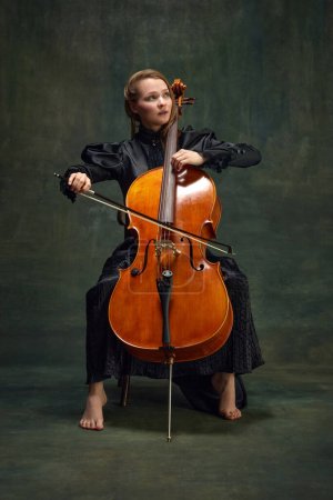 Beautiful woman, talented, passionate cellist looking upward and playing cello against vintage green background. Breathtaking moment. Concept of classical art, retro style, music, inspiration