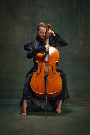 Young woman in black dress, cellist sitting on chair and playing cello against dark green background. Classic music personal therapy. Concept of classical art, retro style, music, inspiration