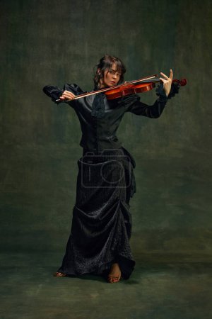 Dynamische Pose einer leidenschaftlichen Musikerin, jungen Frau, Geigerin in schwarzem Gewand, Geige spielend mit intensivem Ausdruck auf altehrwürdigem grünem Hintergrund. Konzept der klassischen Kunst, Retro-Stil, Musik