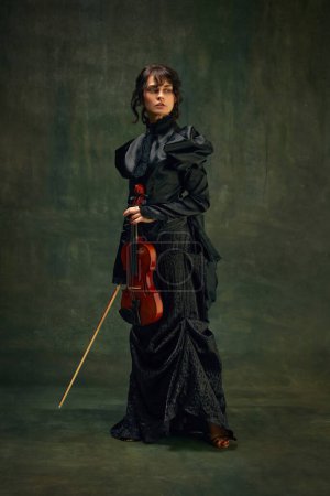 Elégante jeune femme en robe noire, violoniste professionnelle debout avec violon sur fond vert vintage. Musiciens avec performance live. Art classique, style rétro, musique, concept d'inspiration