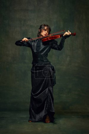 Elegant musician, beautiful young woman in black dress playing violin against dark green vintage background. Symphony. Concept of classical art, retro style, music, inspiration, performance
