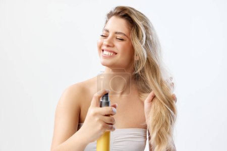 Photo for Smiling woman in strapless top with long blonde hair applying spray against white studio background. Heat protectant products. Concept of beauty, skincare, cosmetics and cosmetology - Royalty Free Image