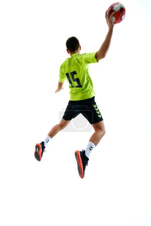 Foto de Imagen de vista trasera dinámica de un joven en uniforme jugando al balonmano en movimiento, saltando con la pelota contra el fondo blanco del estudio. Concepto de deporte profesional, torneo, competición, juego - Imagen libre de derechos