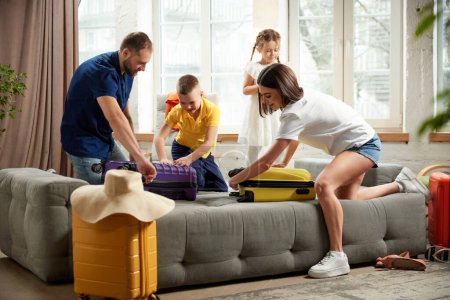 Photo for Man, woman, boy and girl sorting garments for travel, nd packaging bags in bright living room. Joyful mood before trip. Family resort. Concept of tourism, holiday, vacation, relaxation. Ad - Royalty Free Image