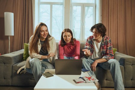 Photo for Live discussion. Young people, men and women, friends sitting on couch at home and booking trip online via laptop. Choosing transport and hotels. Concept of vacation, traveling, booking services - Royalty Free Image