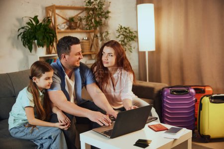 Familia joven, padre, otra y pequeña hija sentada en casa con maletas empacadas y viaje de reserva en línea a través de la computadora portátil. Concepto de vacaciones, viajes, servicios de reserva, en línea
