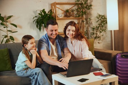 Young family, father, other and little daughter sitting at home with packed suitcases and booking trip online via laptop. Concept of vacation, traveling, booking services, online