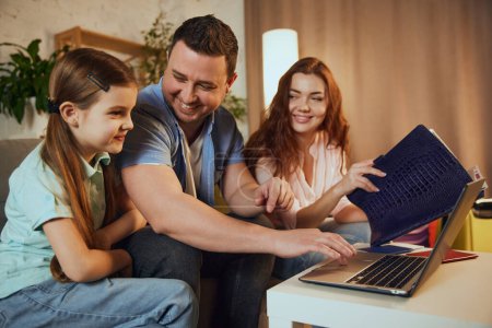 Choosing destination via Internet. Happy family, mother, father and daughter sitting at home, looking on laptop and planning upcoming trip. Concept of vacation, traveling, booking services
