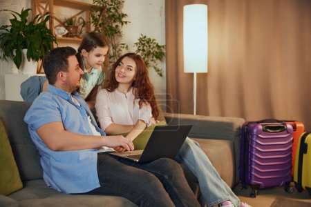 Young family, mother, father and daughter sitting at home with suitcases, planning trip, making online hotel reservation, booking tickets. Concept of vacation, traveling, booking services