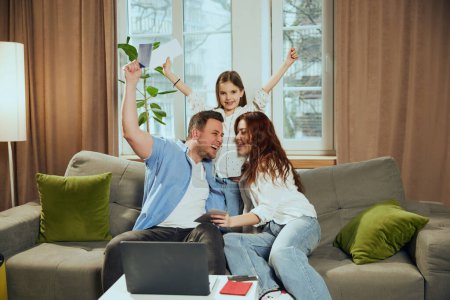 Happy, emotional young family, man, woman and girl buying tickets online via internet booking services, planning trip. Reservation. Concept of vacation, traveling, booking services