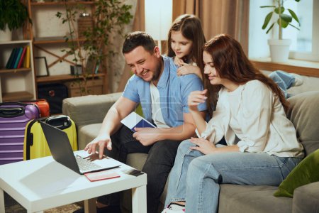Smiling, happy family, mother, father an d daughter sitting on couch at home with suitcases, looking on laptop, making reservation online. Concept of vacation, traveling, booking services