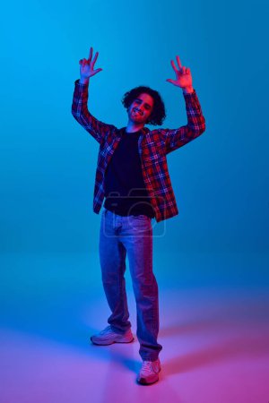 Photo for Dance club, nightlife. Positive young man in casual clothes, checkered shirt and jeans smiling and dancing against blue background in neon light. Concept of youth, human emotions, lifestyle - Royalty Free Image