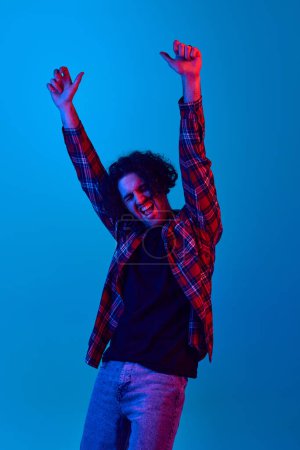 Photo for Happy, excited young man with curly hair, unshaved face expressing positive emotions of success against blue background in neon light. Concept of youth, human emotions, lifestyle. Winner - Royalty Free Image
