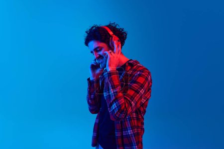 Photo for Young smiling man in casual clothes listening to music in headphones against blue background in neon light. Music vibes, relaxation. Concept of youth, human emotions, lifestyle - Royalty Free Image