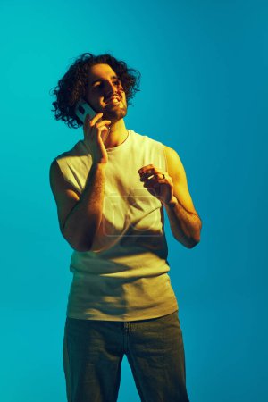 Photo for Young man with curly hair and unshaved face, in casual clothes talking on mobile phone against blue background in neon light. Concept of youth, human emotions, lifestyle - Royalty Free Image