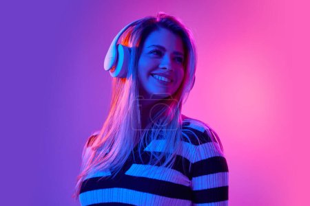 Photo for Positive, attractive young woman smiling, wearing headphones and listening to music against pink studio background in neon light. Concept of human emotions, youth, fashion, expression - Royalty Free Image