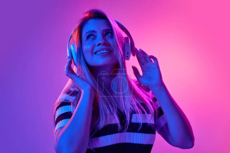 Foto de Retrato en gel de una hermosa mujer joven escuchando música en auriculares con expresión de ensueño contra fondo de estudio rosa en luz de neón. Concepto de emociones humanas, juventud, moda, expresión - Imagen libre de derechos