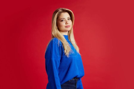 Foto de Retrato de una joven atractiva con el pelo rubio, vistiendo suéter azul, mirando a la cámara contra el fondo rojo del estudio. Bienestar. Concepto de emociones humanas, juventud, moda, expresión - Imagen libre de derechos
