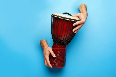 Foto de Las manos masculinas sobresalen del fondo azul y juegan al djembe. Cartel del festival de música mundial con diversos instrumentos tradicionales. Concepto de música, instrumentos, arte, hobby, festival, performance - Imagen libre de derechos