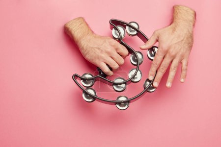 Male hands sticking out pink background and playing tambourine. Latin studio promotional image showcasing traditional instruments. Concept of music, instruments, art, hobby, festival, performance