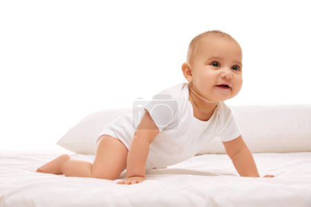 Foto de Adorable niña pequeña, niño pequeño en ropa blanca, arrastrándose sobre malos con almohadas sobre fondo blanco. Ocio, movimientos. Concepto de infancia, familia, cuidado, maternidad, infancia, salud - Imagen libre de derechos