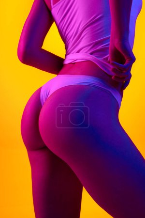 Téléchargez les photos : Image recadrée d'une femme mince avec ajustement, les fesses lisses sur fond orange dans la lumière au néon violet. Soin anti-cellulite. Concept de soins corporels et de santé, beauté féminine, bien-être - en image libre de droit