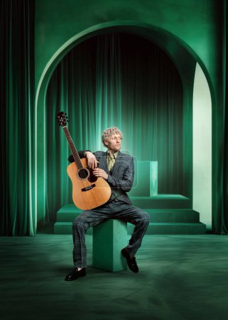 Man in suit with calm expression, musician sitting with guitar against green luxurious stage. Promotional material for live concert. Concept of music, performance, art, talent show, inspiration