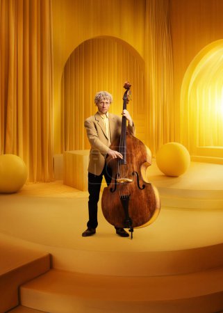 Photo for Male musician in suit with double bass in monochromatic yellow luxurious room. Elegance and classical taste. Concept of music, performance, art, talent show, inspiration. Poster - Royalty Free Image