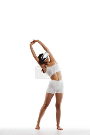 Foto de Imagen completa de chica joven con cuerpo delgado, con gafas VR y entrenamiento, haciendo estiramiento sobre fondo blanco del estudio. Concepto de cuerpo y salud, deporte, belleza femenina, bienestar - Imagen libre de derechos