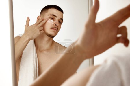 Téléchargez les photos : Jeune homme dans la salle de bain regardant dans le miroir et hydratant visage après le rasage. Concentration sélective. Concept de soins de beauté, santé masculine, soins du corps, soins spa, hygiène. - en image libre de droit