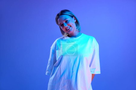 Foto de Retrato en gel de la hermosa joven en camiseta blanca de pie, posando con sonrisa sobre fondo de estudio azul en luz de neón. Concepto de juventud, emociones humanas, moda casual - Imagen libre de derechos