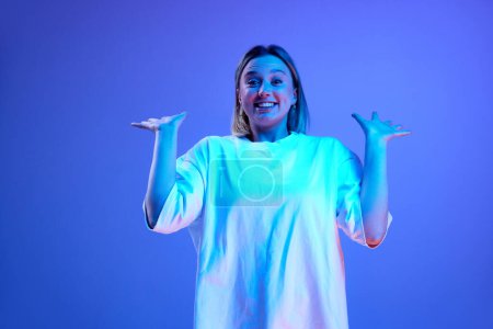 Téléchargez les photos : Portrait en gel d'une jeune fille heureuse et excitée en t-shirt blanc posant avec sourire sur fond de studio bleu sous la lumière du néon. Surprise. Concept de jeunesse, émotions humaines, mode décontractée - en image libre de droit