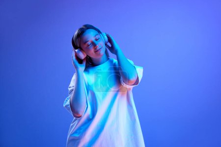Foto de Mujer joven positiva en camiseta blanca casual escuchando música en auriculares contra fondo de estudio azul en luz de neón. Concepto de juventud, emociones humanas, moda casual - Imagen libre de derechos
